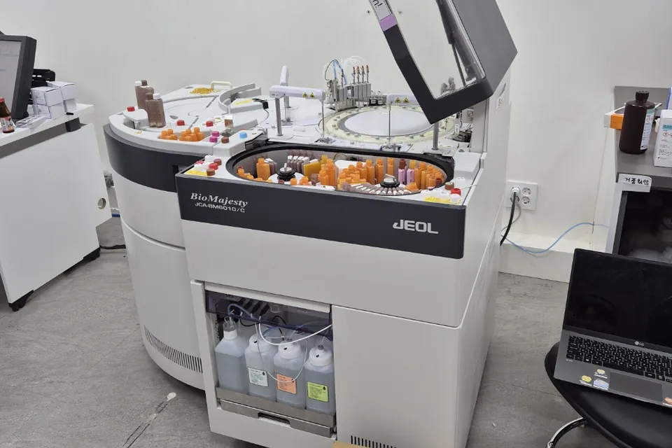 서울 소재 병원 검사실에 설치된 JEOL JCA-BM6010/C 생화학분석기가 덮개를 연 채 시약병이 꽂힌 원형 트레이를 드러내고 있습니다. 오른쪽 의자 위에 노트북이 있습니다.