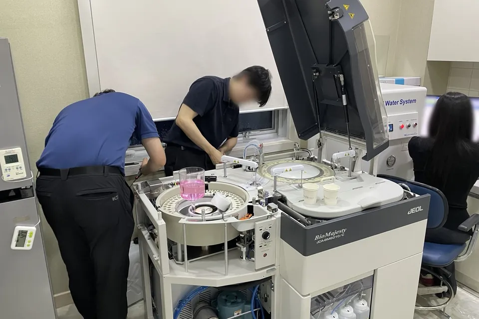 울산 소재 내과의원 검사실에서 두 사람이 덮개를 연 JEOL JCA-BM6010/C 생화학분석기를 설치하고 있고, 오른쪽에 한 사람이 앉아 있습니다. 인물 얼굴은 흐리게 처리했습니다.