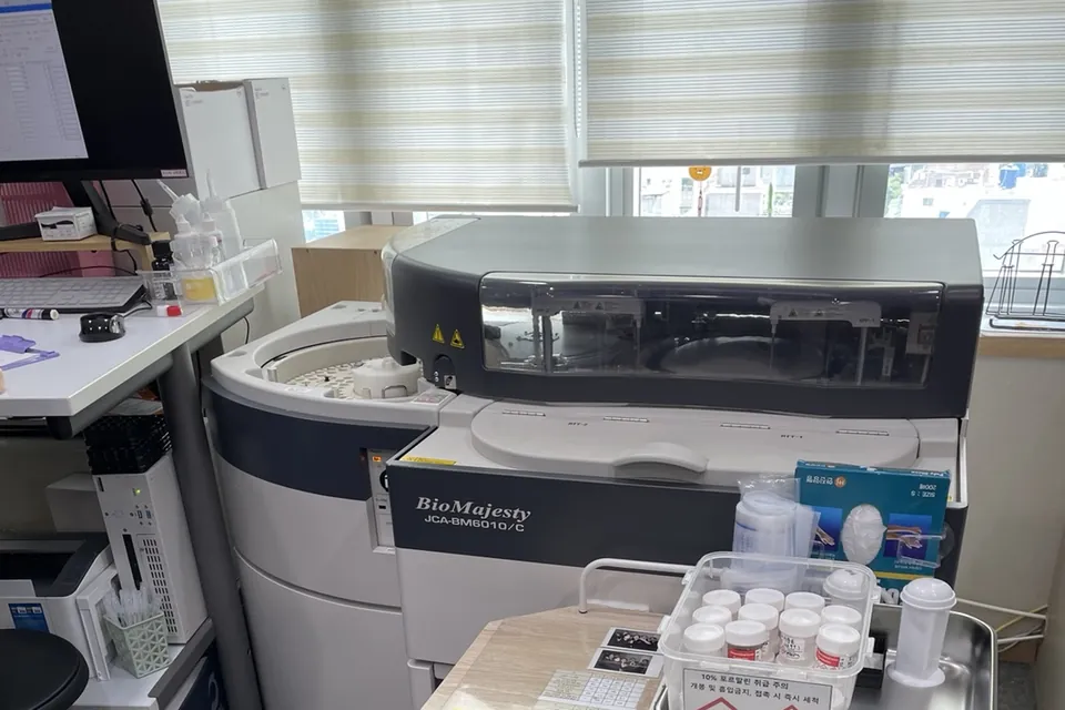 창원 소재 병원 검사실 창가에 설치된 JEOL JCA-BM6010/C 생화학분석기. 앞쪽 카트에 검체 용기 상자와 장갑 상자가 놓여 있습니다.